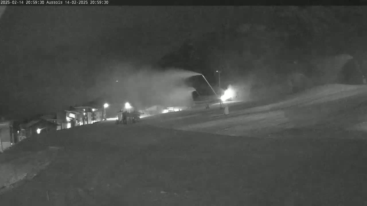 At night, a snow groomer sprays snow onto a slope near buildings under a dark, clear sky.