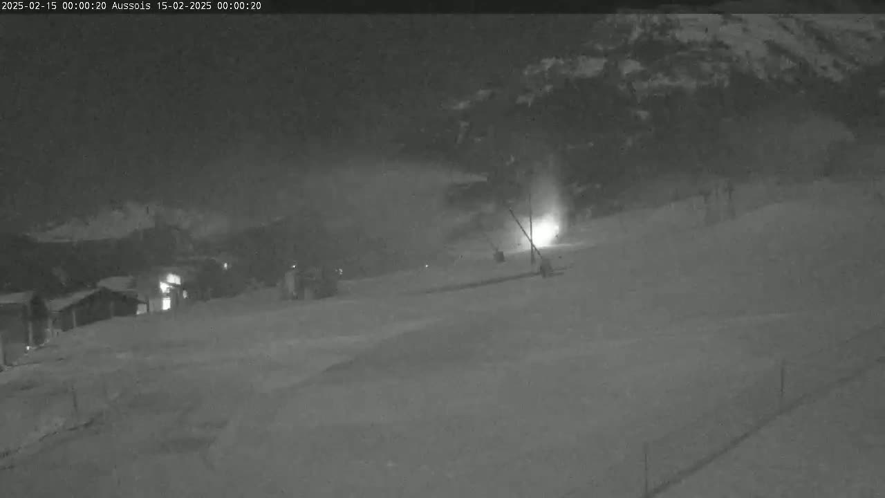 A nighttime, snowy scene shows a lightly-snowing ski slope with a few lights visible in the distance.