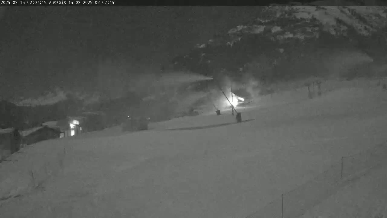 A nighttime snow scene shows a snow-covered slope with some structures and a bright light source in the mid-ground, possibly snowmaking equipment.