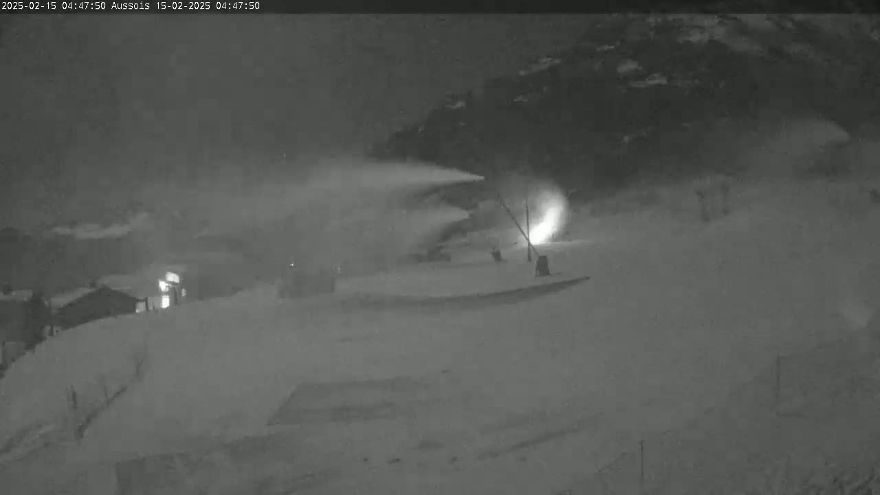 A snowmaking machine is operating at night in a snowy, outdoor setting, near some buildings.
