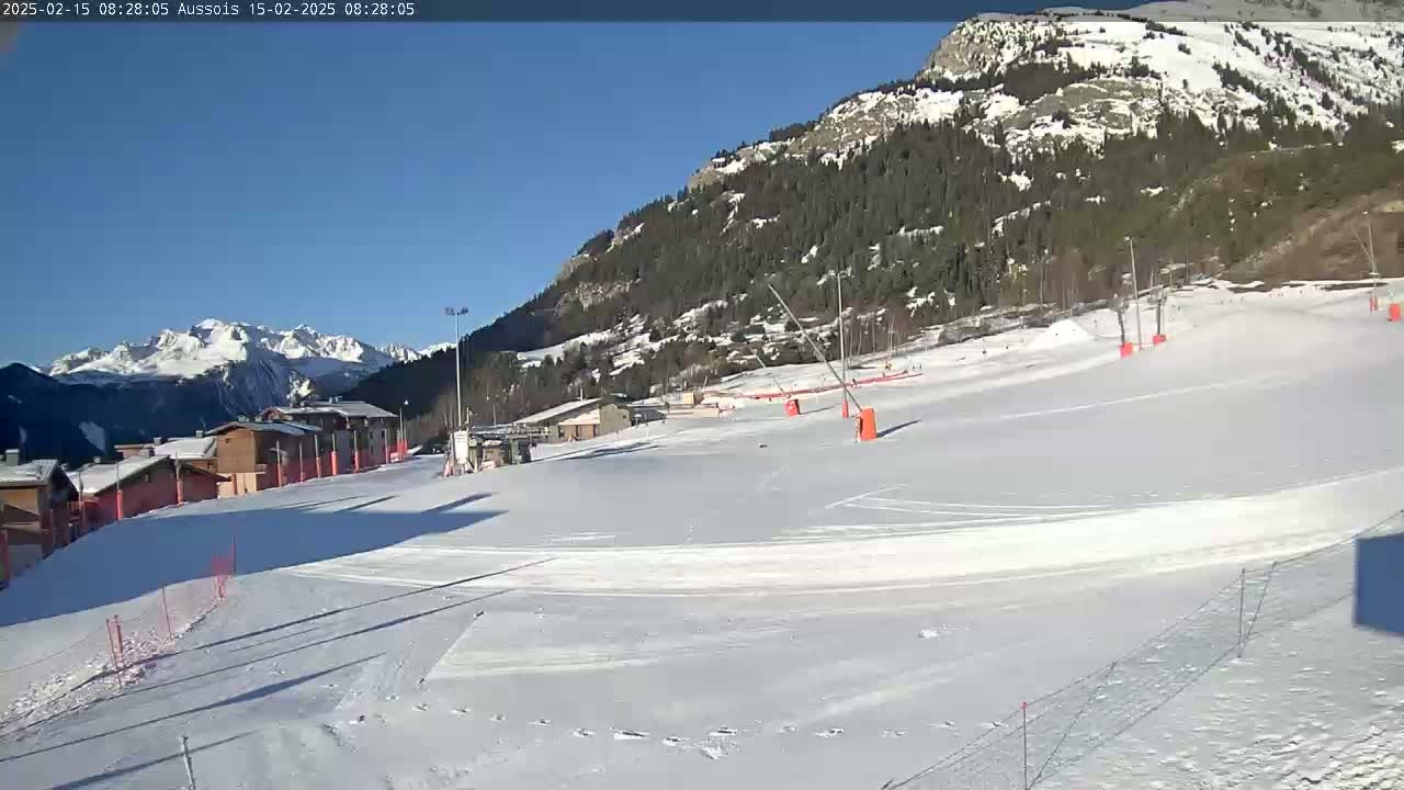 A snow-covered ski slope is nestled beneath a sunny, clear sky, with mountains and buildings in the background.