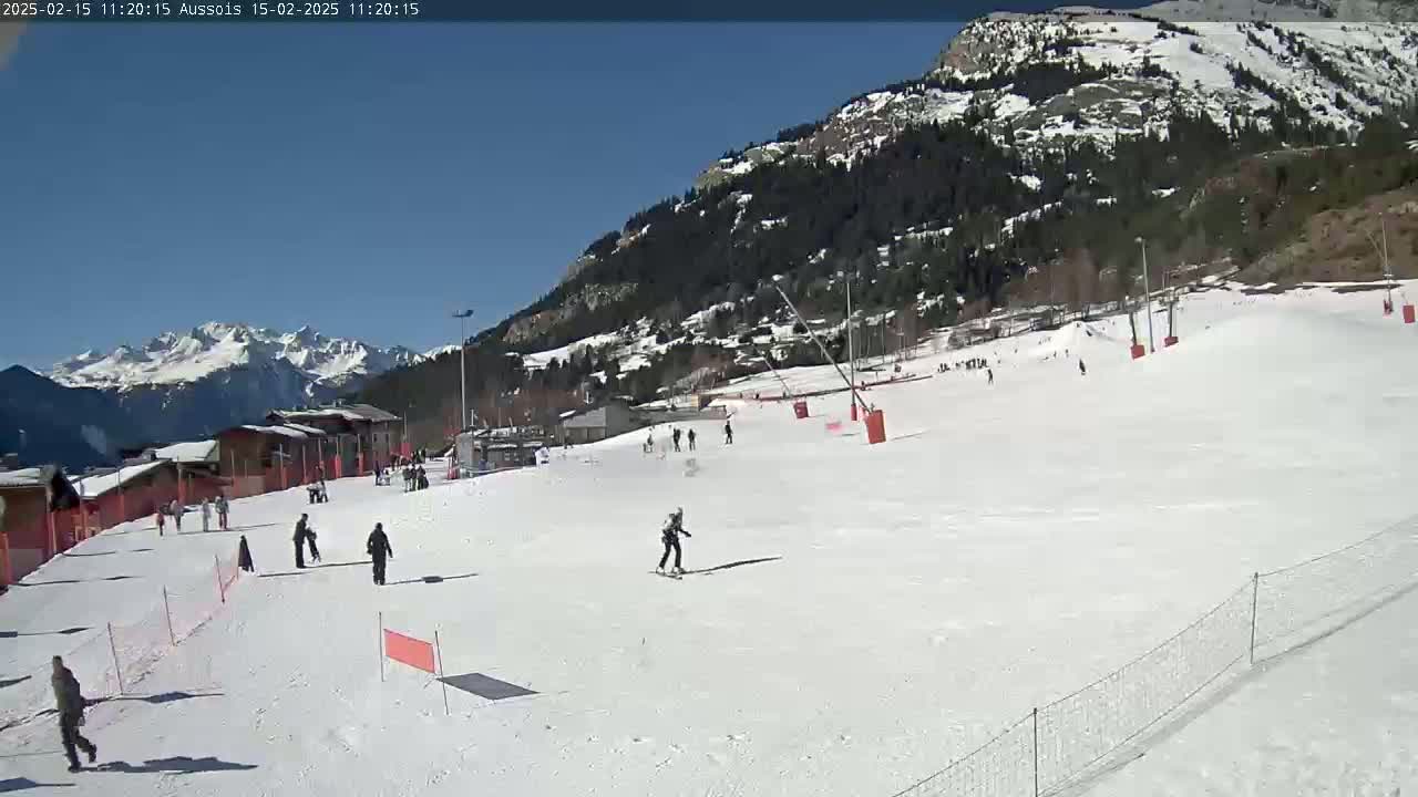 A sunny day on a snow-covered ski slope with several skiers and snowboarders, backed by a mountain range and a village.