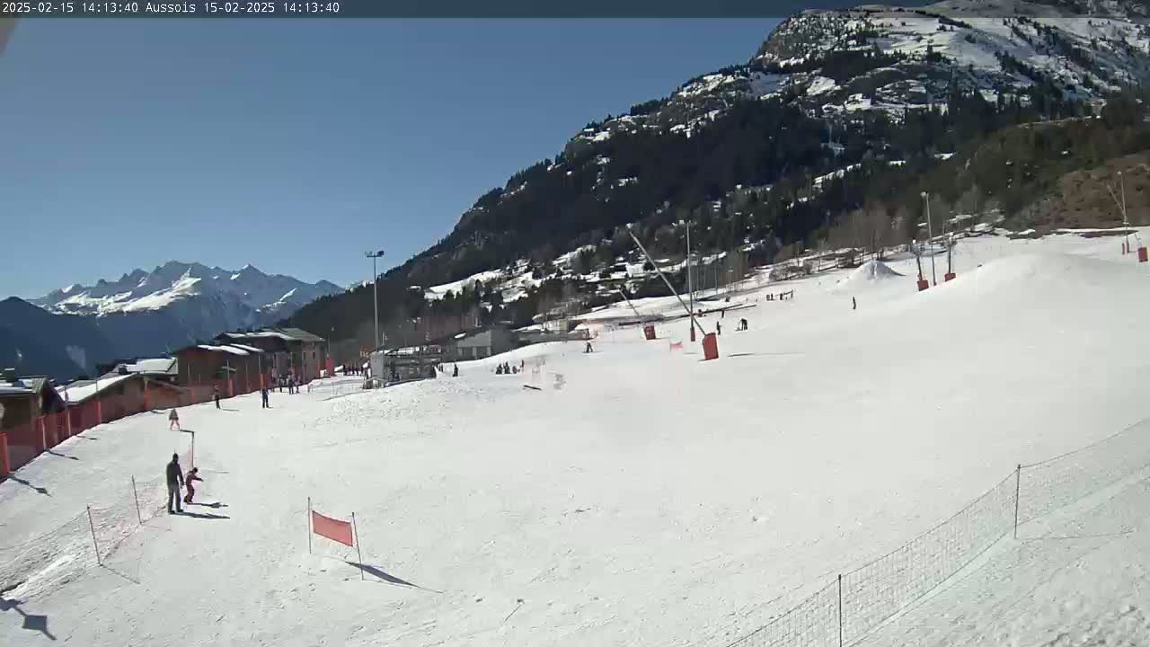 A sunny day at a snow-covered ski slope with several people skiing and snowboarding, set against a backdrop of mountains.