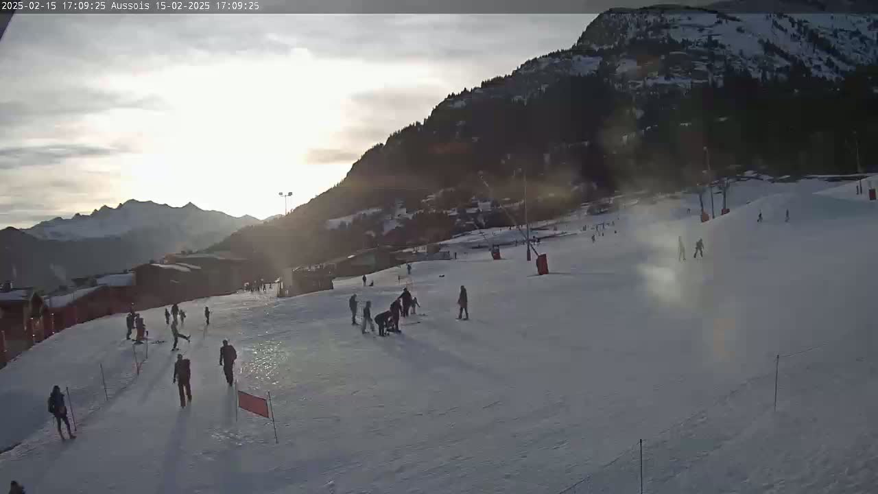 A snowy ski slope with several people skiing and snowboarding, situated against a mountainous backdrop under a partly cloudy sunset sky.