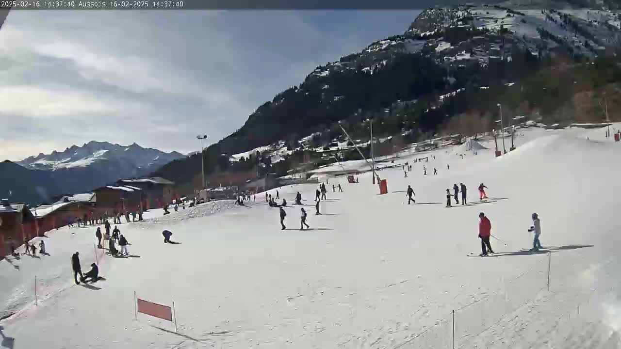 A snow-covered ski slope with several skiers and snowboarders, some buildings, and a mountain backdrop under a mostly sunny sky.