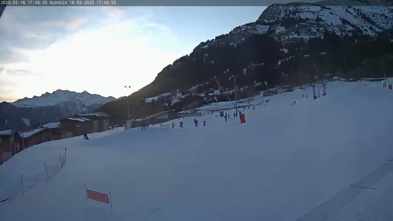 A mostly sunny, snow-covered ski slope with several skiers and snowboarders, situated beneath a mountain at dusk.