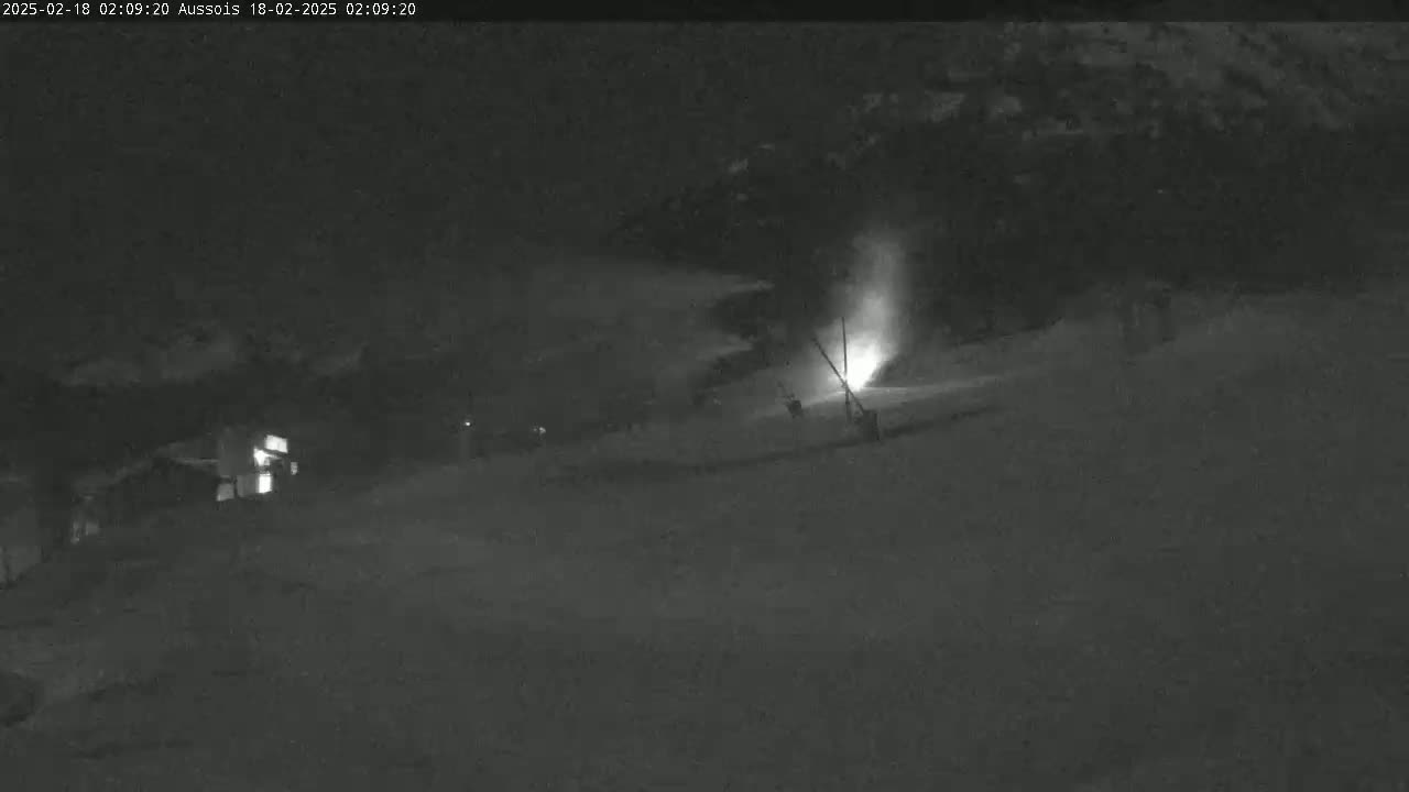 A nighttime, snow-covered scene shows a faintly lit area with what appears to be a snow-making machine emitting a plume of snow, near a dark building.