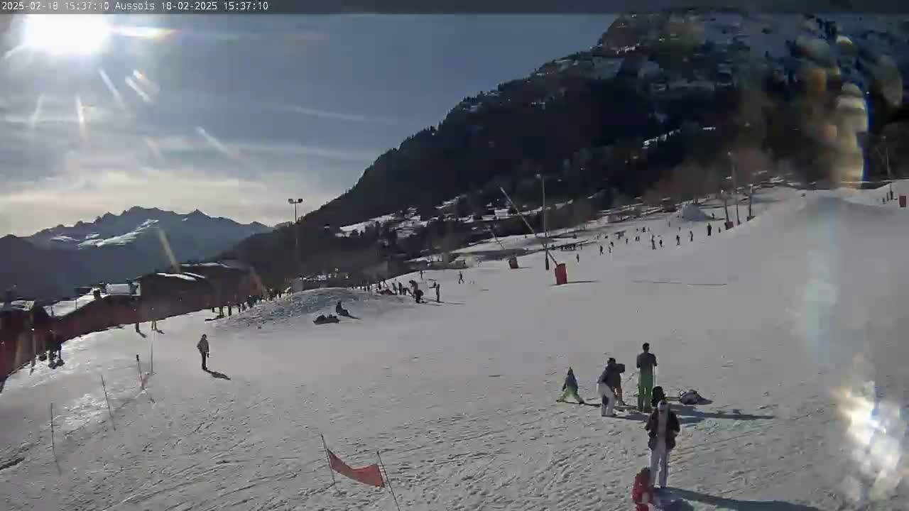 A sunny day on a snow-covered ski slope with several people skiing and snowboarding, mountains in the background.