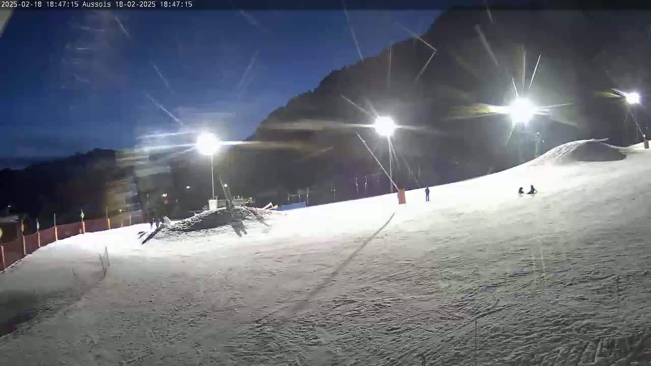 A snow-covered slope at night, illuminated by bright lights, with a few people visible.