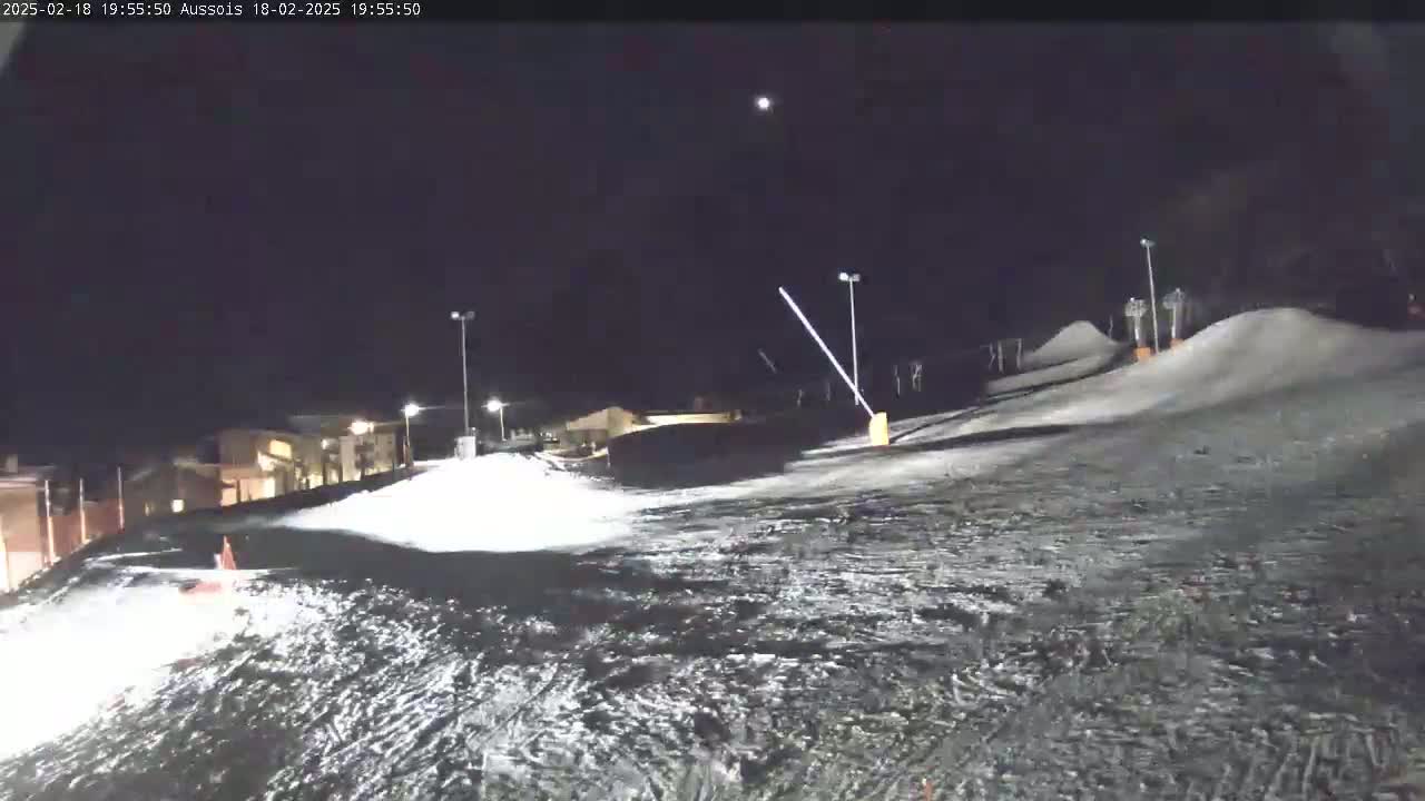 A snow-covered slope at night is illuminated by several lights, with a building visible in the background under a clear, dark sky.