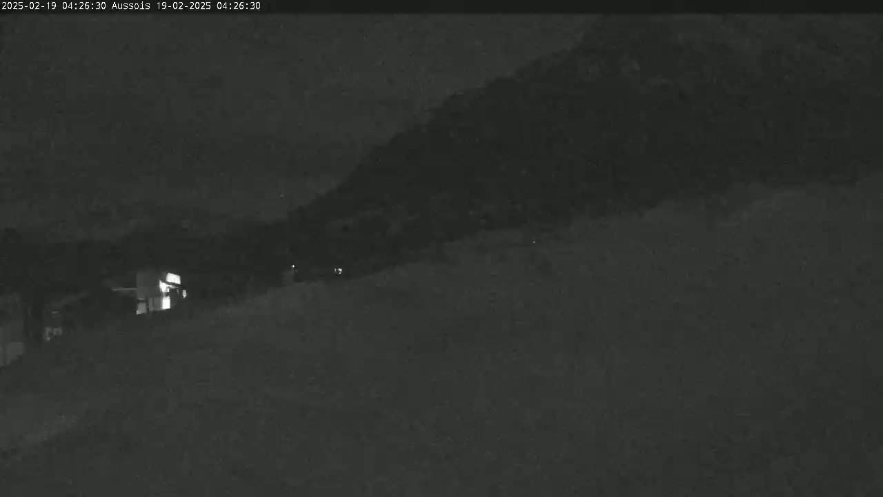 A nighttime, low-light view shows a dark, hilly landscape with a single lit building visible in the lower left.