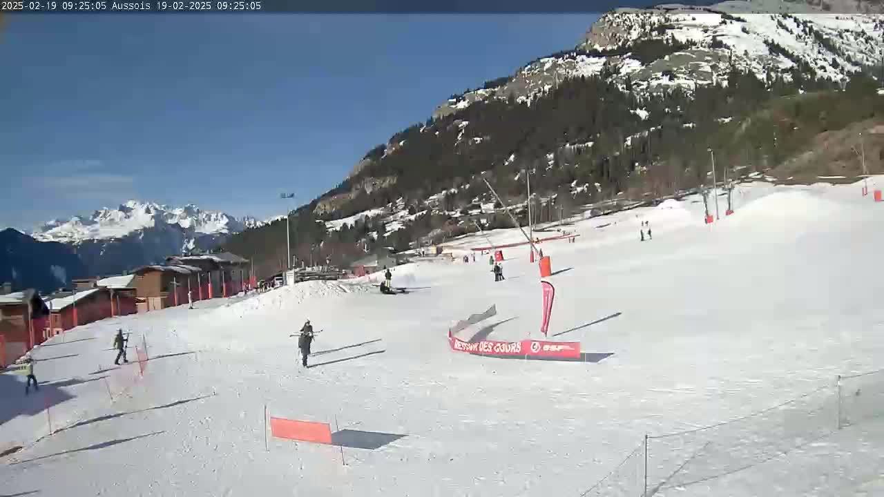 A sunny snow-covered ski slope with several skiers and snowboarders, buildings, and mountains in the background.