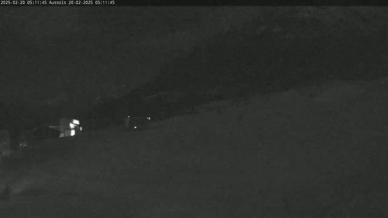A nighttime, low-light image shows a dark, indistinct landscape with a few faint lights visible in the mid-ground.