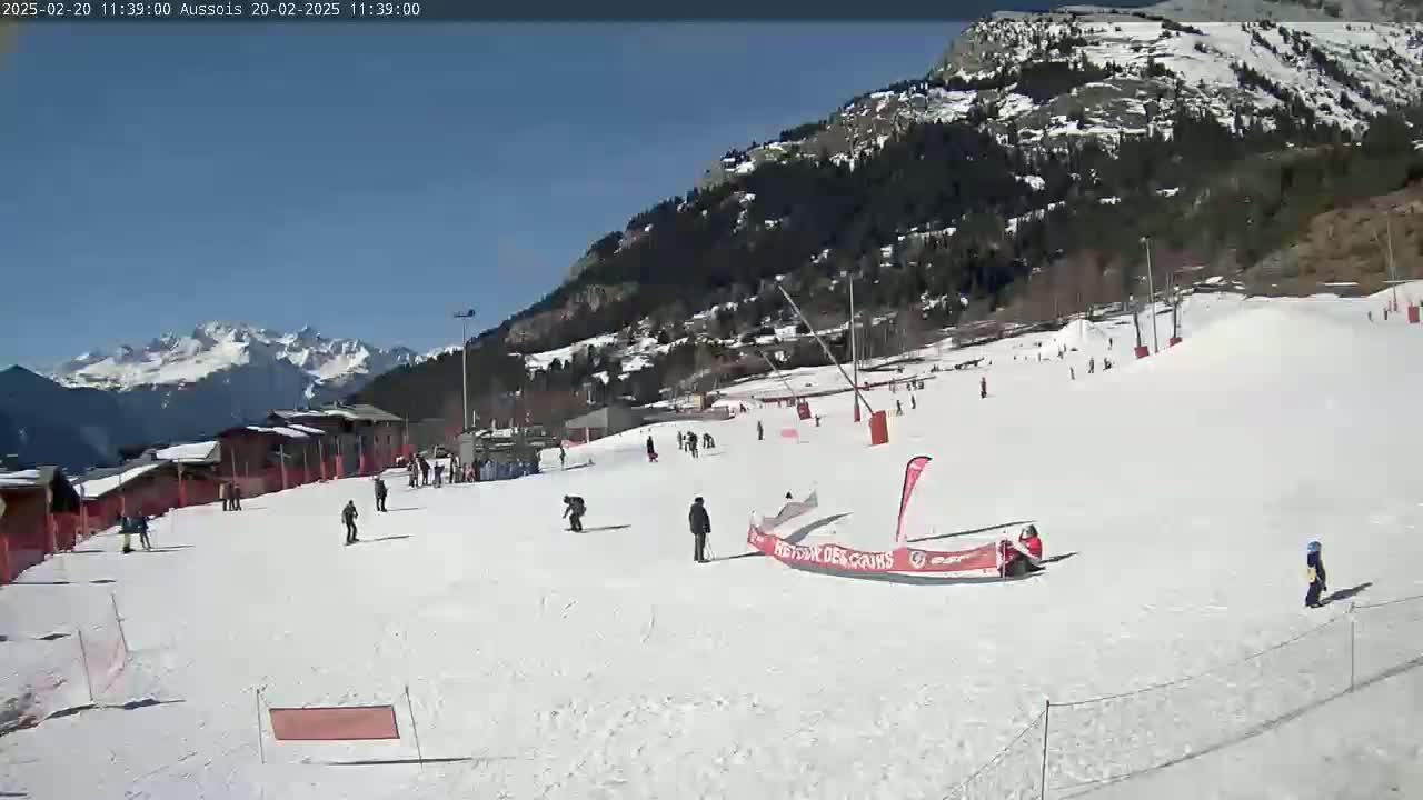 A sunny day at a snow-covered ski resort shows skiers and snowboarders on a slope against a backdrop of mountains.