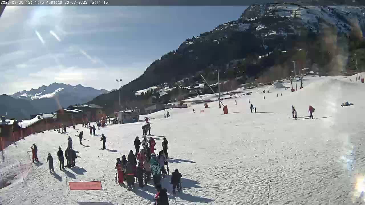 A sunny day on a snow-covered ski slope with several people skiing and snowboarding, surrounded by mountains and buildings.