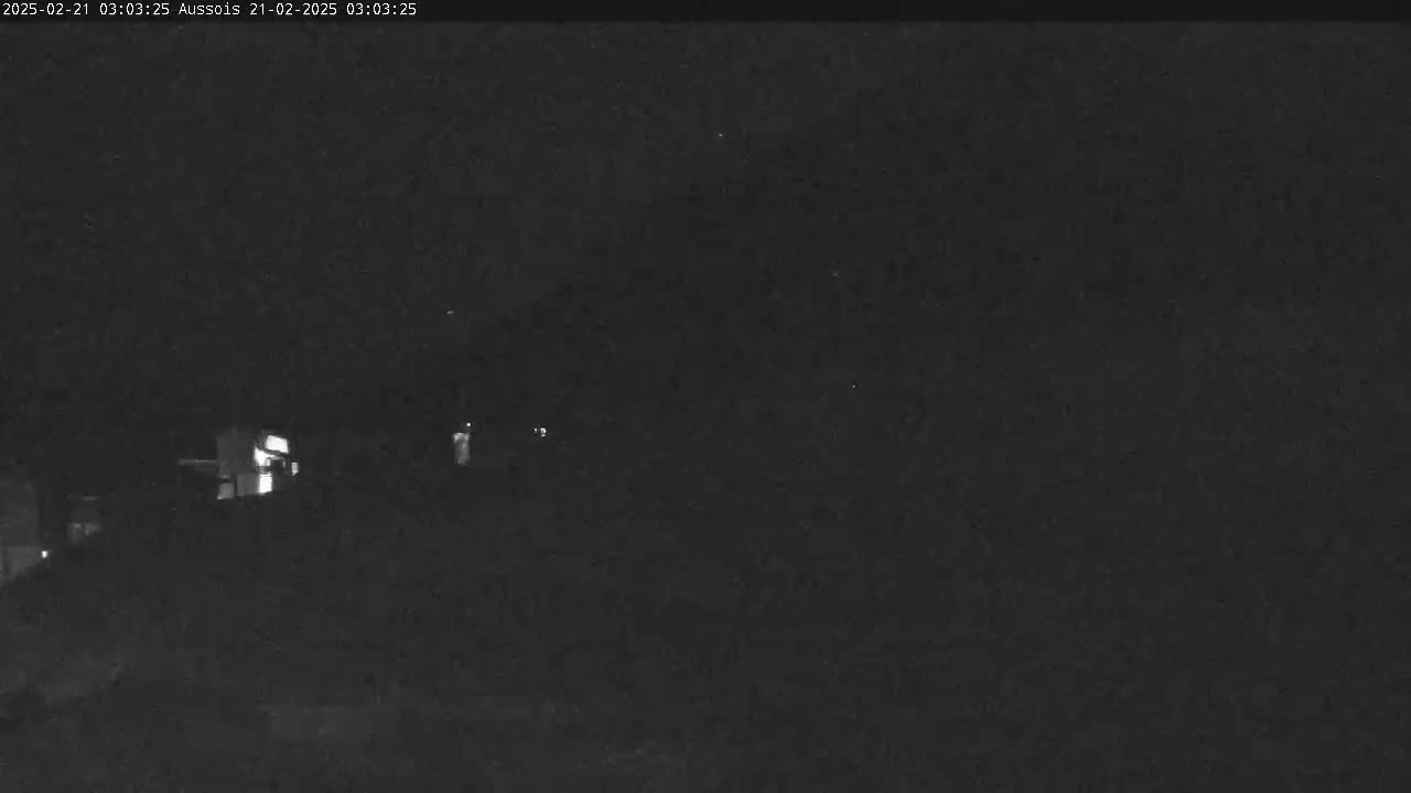 A low-light, nighttime image shows a dark landscape with faintly illuminated structures in the distance.