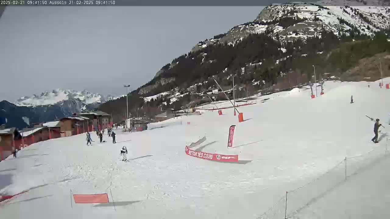 A snowy ski slope with several skiers and snowboarders, backed by a mountain range under a mostly clear sky.