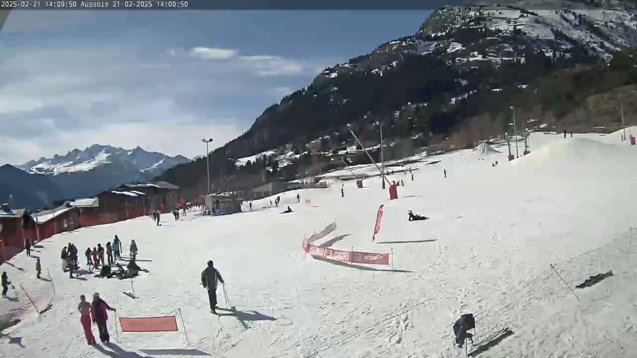 A sunny day at a snow-covered ski resort shows several people skiing and snowboarding on a wide slope, with mountains in the background.