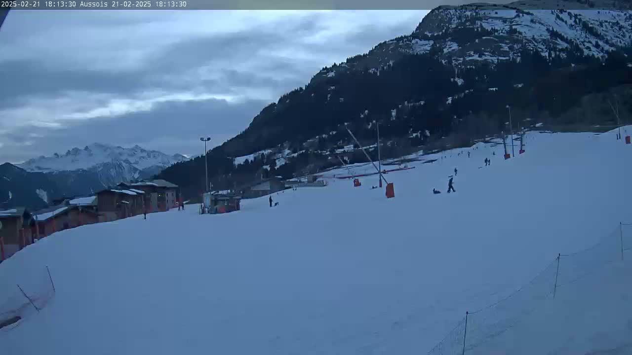 A snow-covered ski slope with several people skiing or snowboarding, under a cloudy twilight sky, backed by a mountain range.