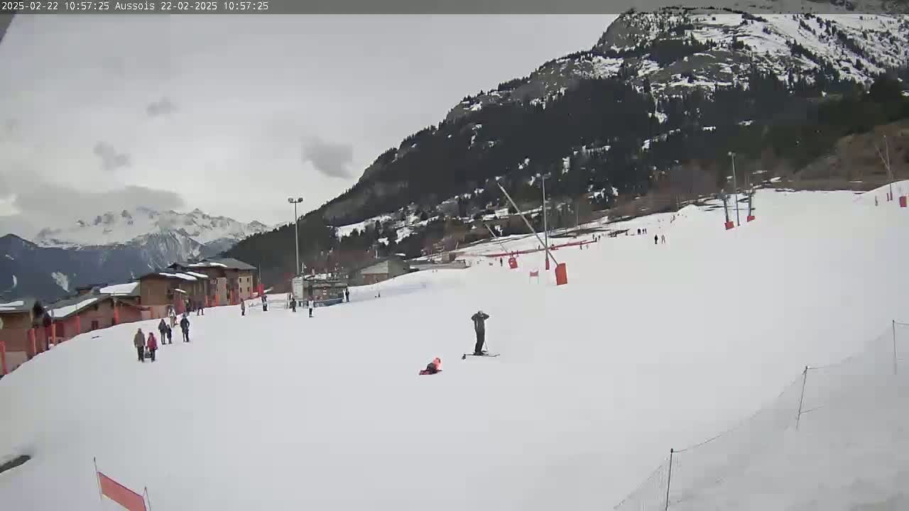 A mostly cloudy day reveals a snow-covered ski slope with several skiers and snowboarders, backed by a mountain range and buildings.