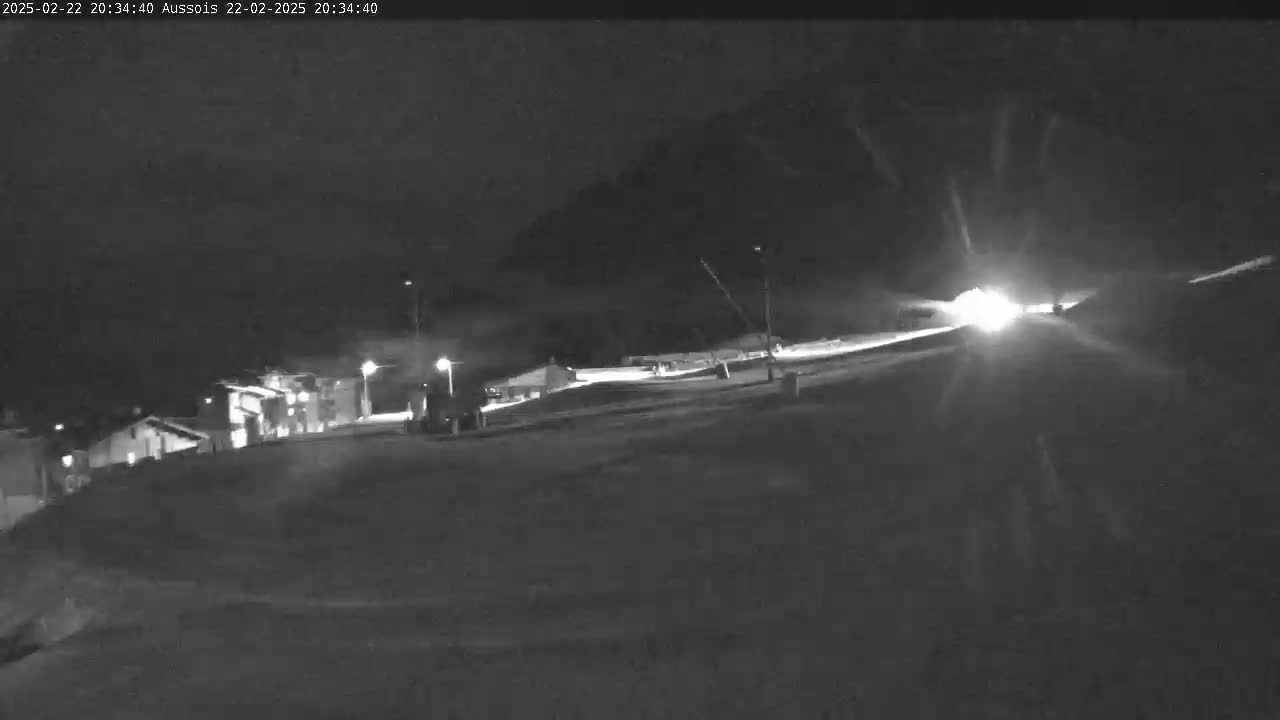 A nighttime, snow-covered slope is illuminated by headlights of vehicles traveling uphill towards a building complex.