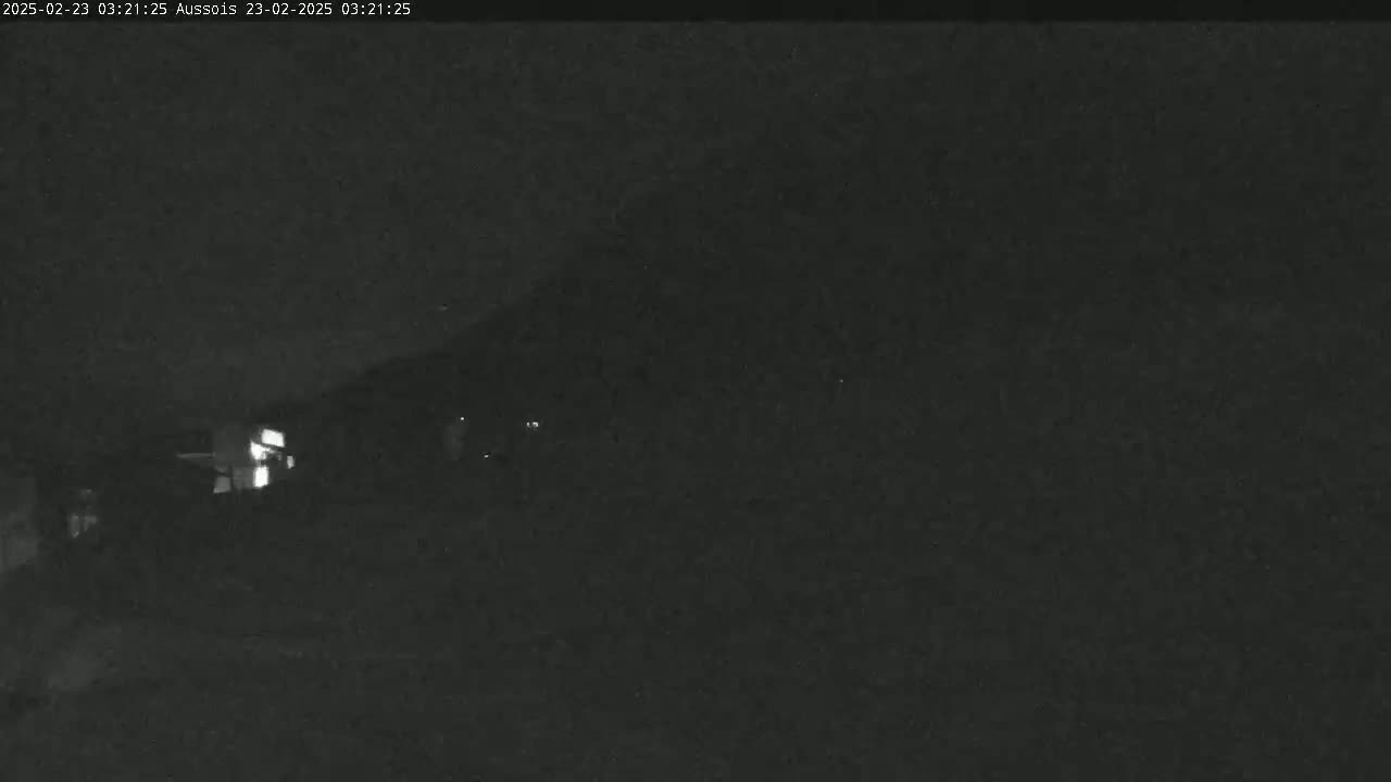 A nighttime, low-light image shows a dark, mostly indistinct scene with a dimly lit building partially visible in the lower left.