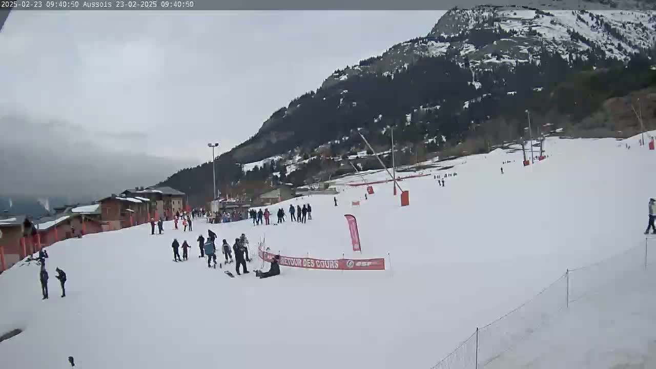 A snowy ski slope with several skiers and snowboarders, ski lifts, and buildings in the background under an overcast sky.