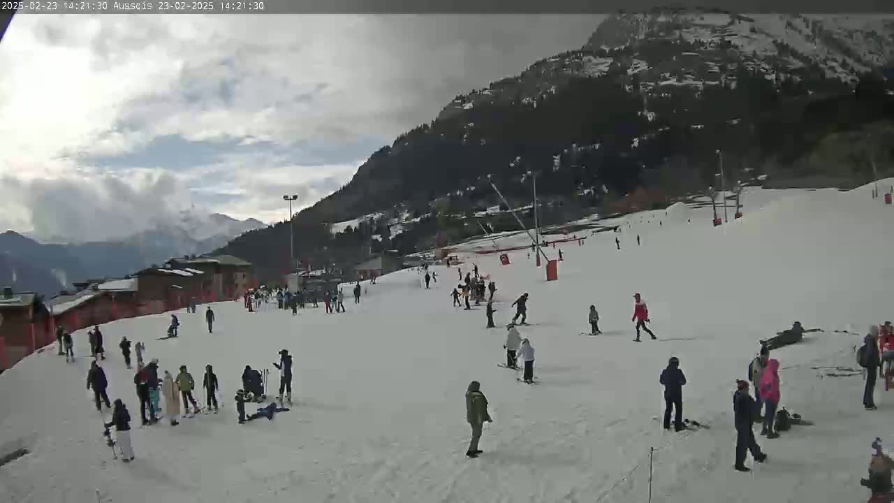 A snowy ski slope with several skiers and snowboarders, under a cloudy sky, is situated at the base of a mountain.