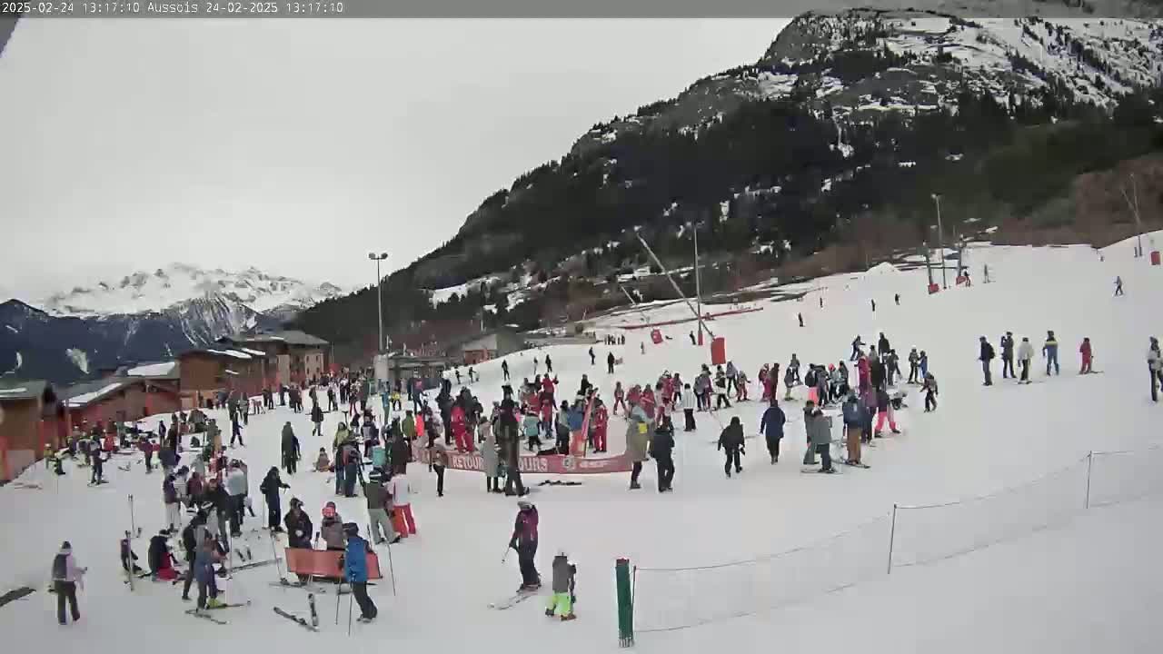 A snowy mountain slope with numerous skiers and snowboarders, many gathered in groups, under an overcast sky.