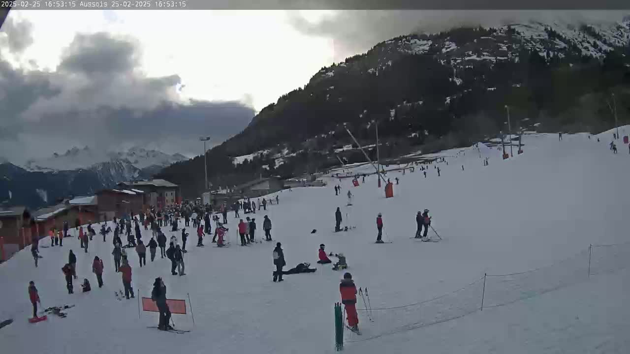 A snow-covered ski slope with skiers and snowboarders, some buildings, and a mountain backdrop under a cloudy sky.