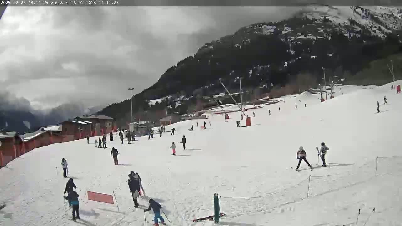 A snowy ski slope with skiers and snowboarders, under a cloudy sky, is situated in front of a mountain covered in dark evergreen trees.