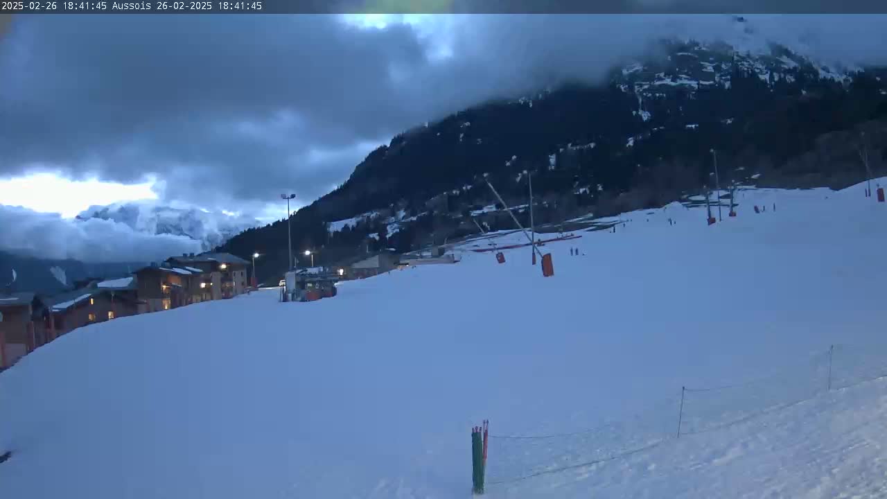 A snow-covered ski slope with buildings and ski lifts is visible under a cloudy, twilight sky.