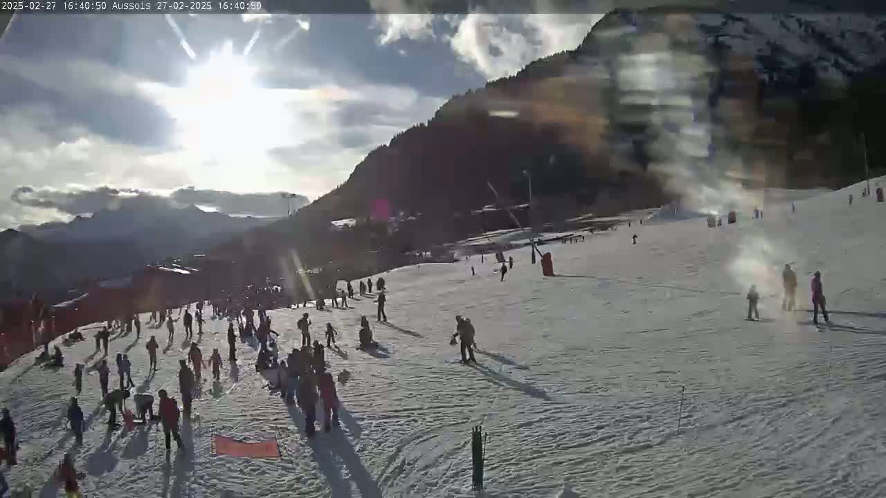 A sunny day on a snow-covered ski slope with numerous skiers and snowboarders, mountains in the background.