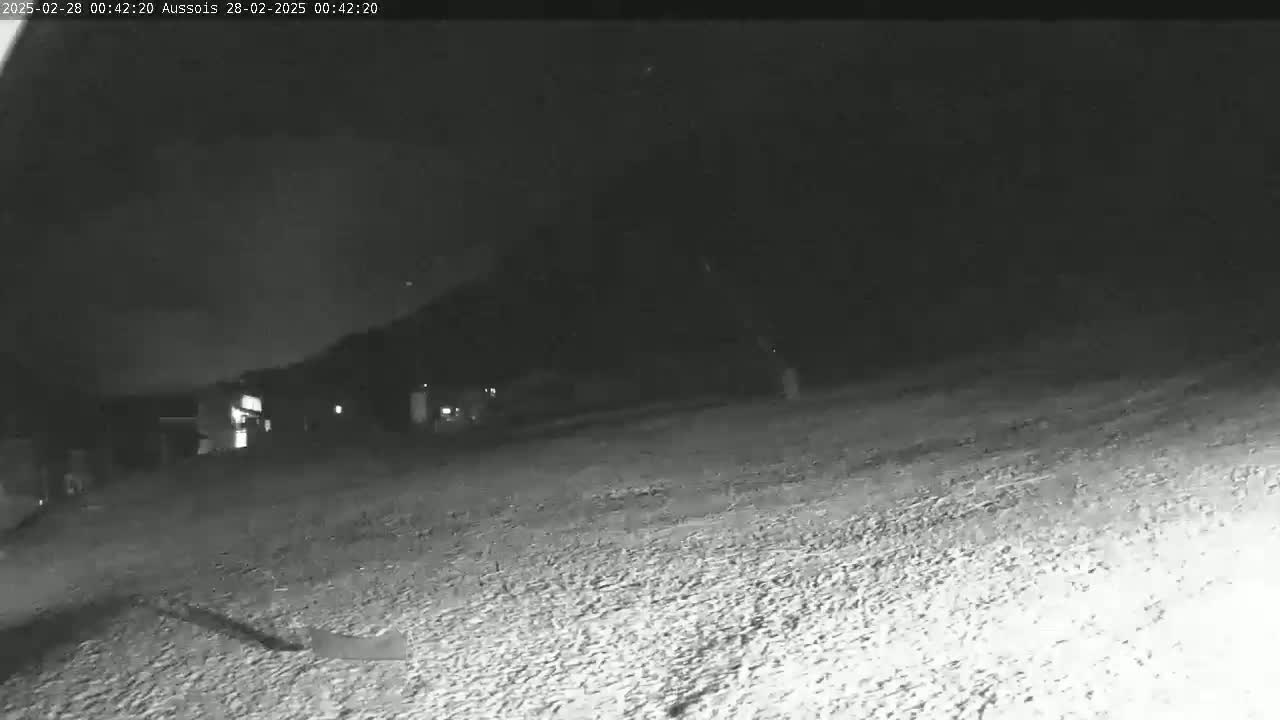 A low-light, nighttime image shows a mostly empty, snow-covered ground with dimly lit buildings in the distance.