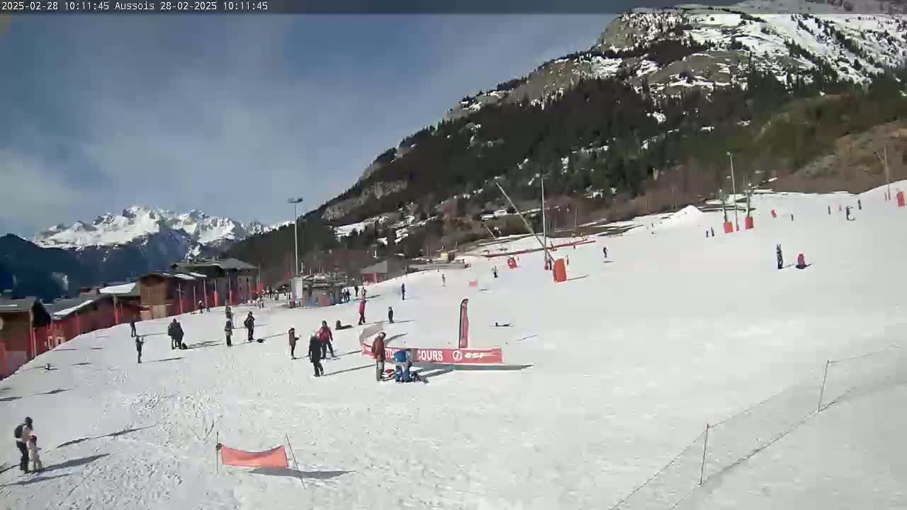 A sunny day on a snow-covered ski slope with several people skiing and snowboarding, with mountains in the background.