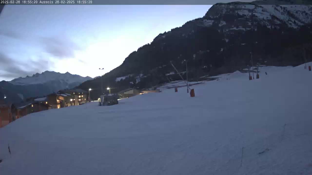 A snow-covered ski slope at dusk, with a mountain in the background and some buildings with lights on in the distance.