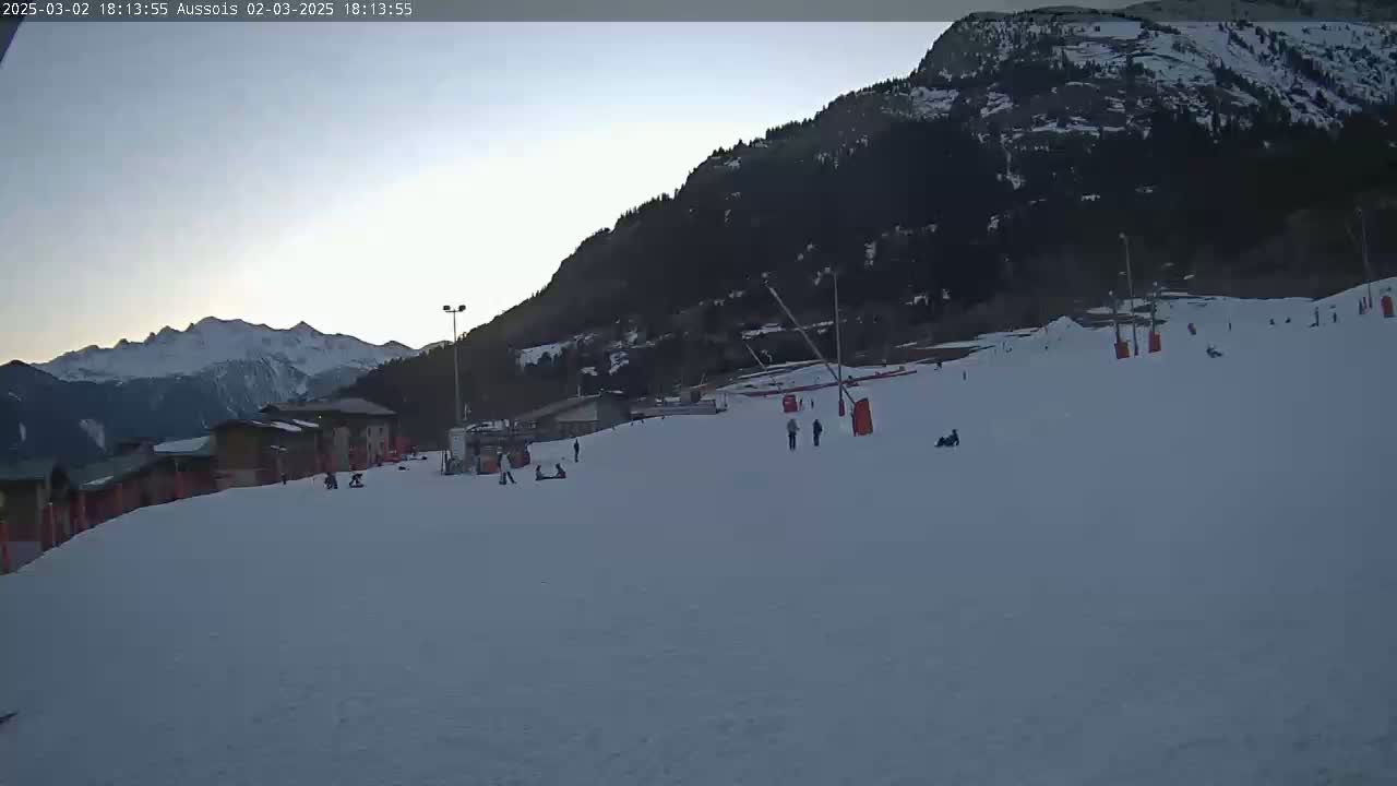 A snow-covered ski slope with a few skiers and snowboarders, situated below a mountain range under a twilight sky.