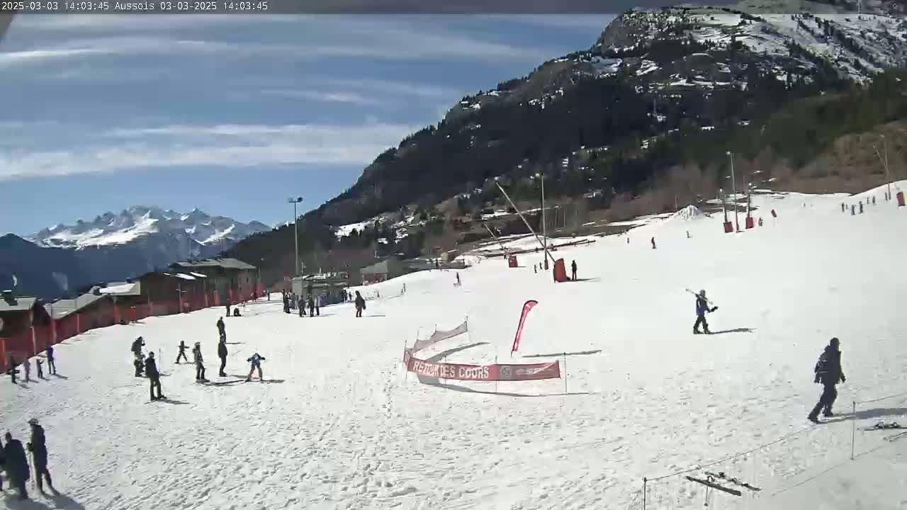 A sunny day at a snow-covered ski slope with several people skiing and snowboarding, mountains in the background.