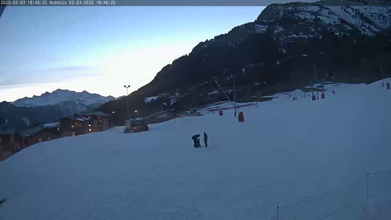 A snow-covered ski slope at dusk, with a mountain range in the background and a few people near a building.