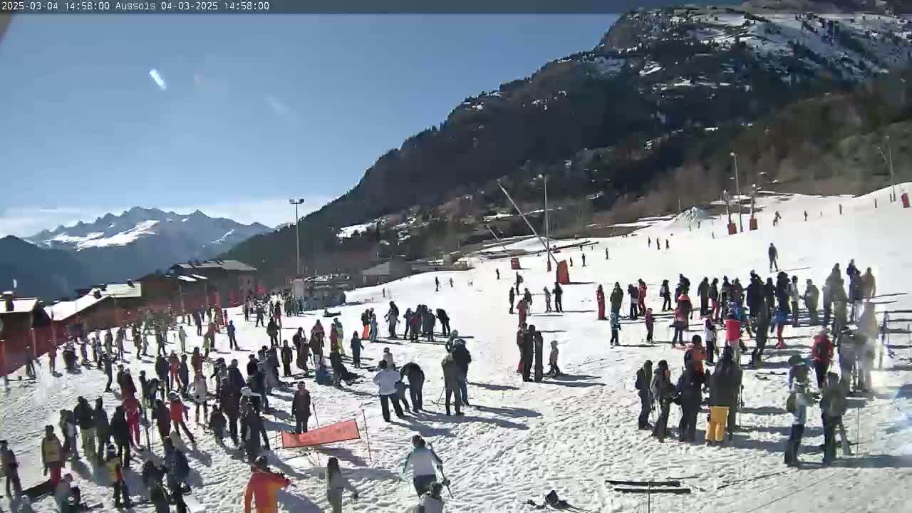 A sunny day at a snow-covered ski resort shows numerous people skiing and gathering on the slopes, with mountains in the background.