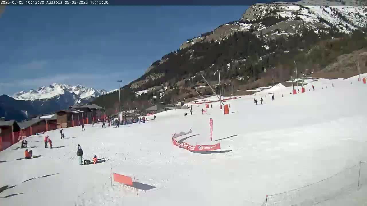 A snow-covered ski slope with several people skiing and snowboarding, situated against a backdrop of mountains under a clear blue sky.