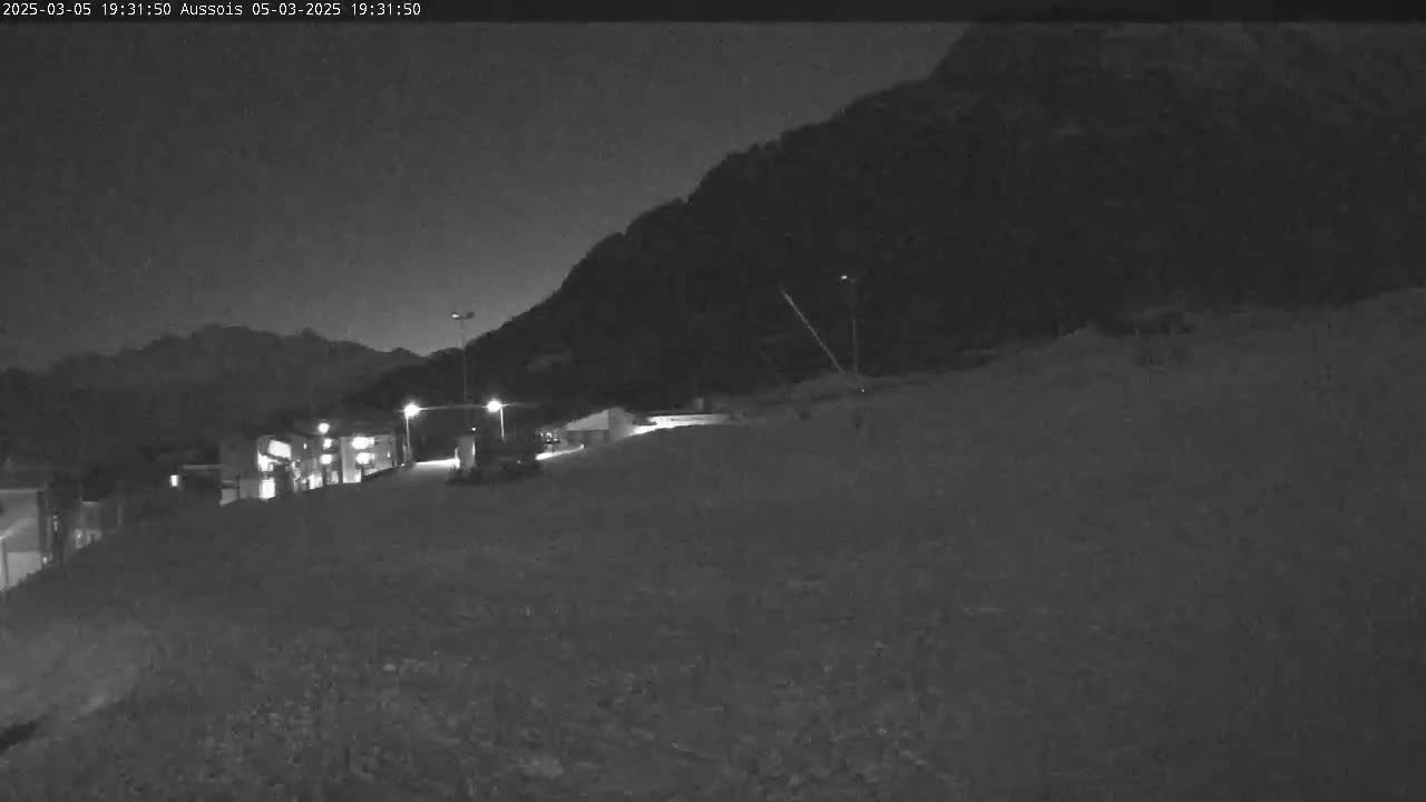 A nighttime view of a snow-covered area with a few buildings and lights visible against a dark mountain backdrop.