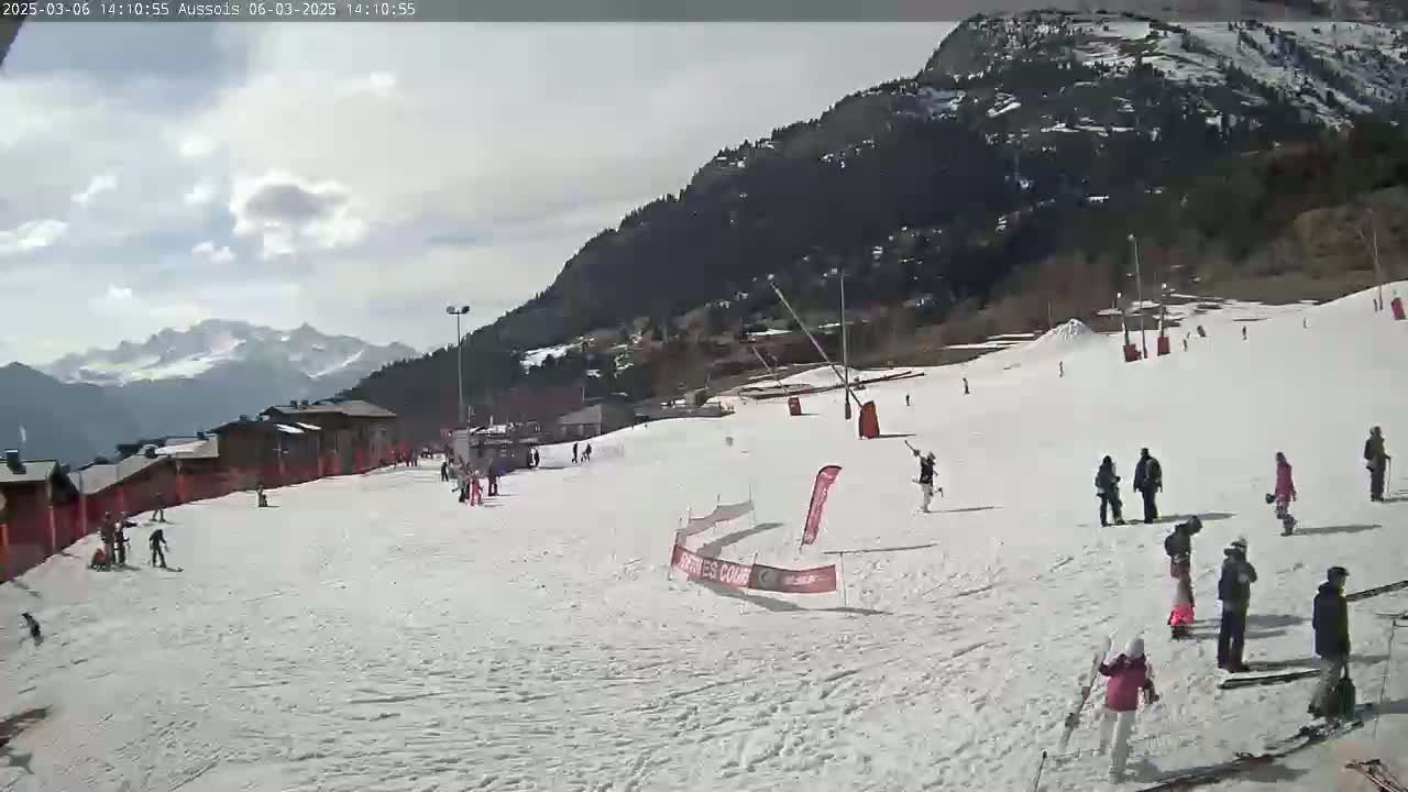 A snowy ski slope with several skiers and snowboarders is visible, with a mountain range in the background under a partly cloudy sky.