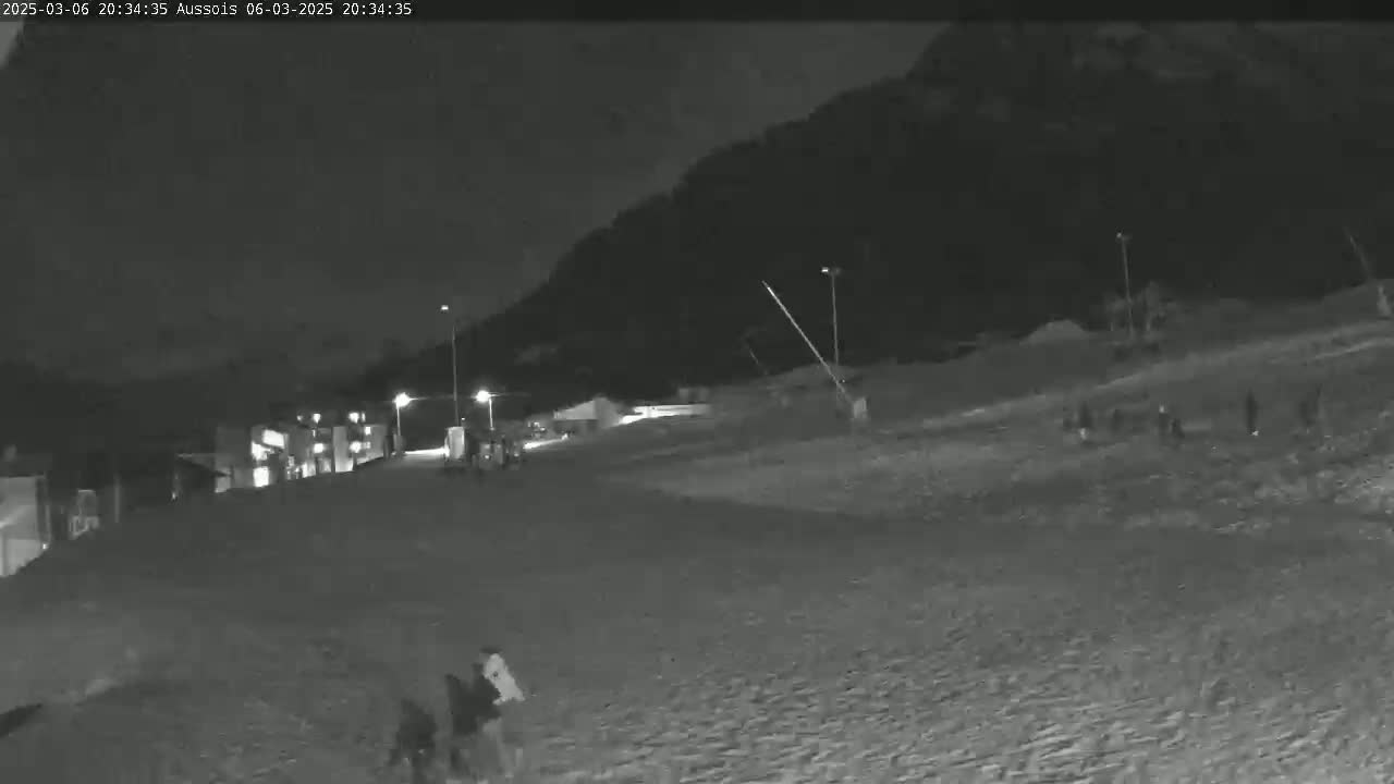 A nighttime, low-light grayscale image shows a sloped area with several people walking, buildings in the background, and a mountain in the distance.