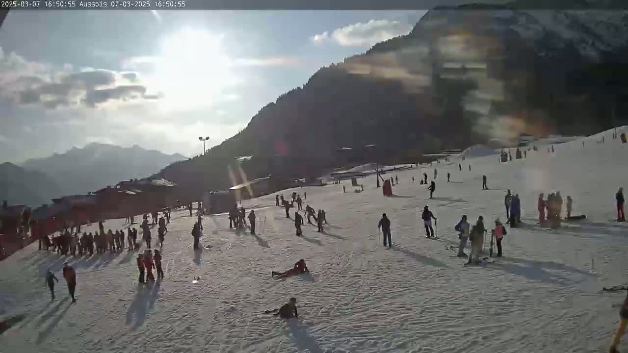 A sunny day with partly cloudy skies shows many people skiing and snowboarding on a snow-covered mountain slope.