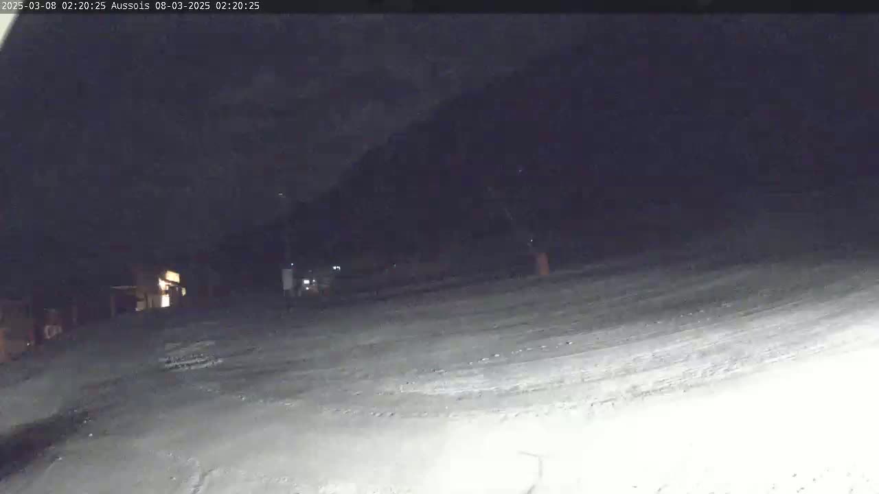 A snow-covered area is visible at night, with a dark mountain in the background and a few lights from buildings in the distance.