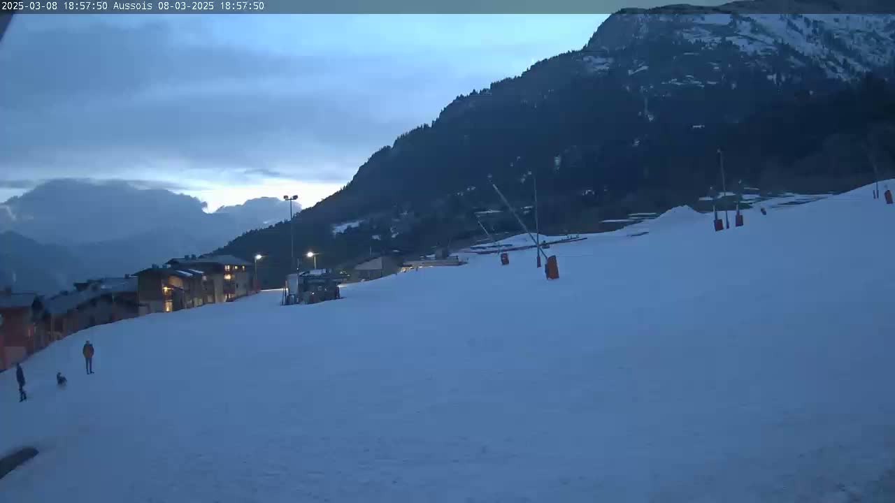 A snowy ski slope at dusk, with a few people walking and a mountain in the background.