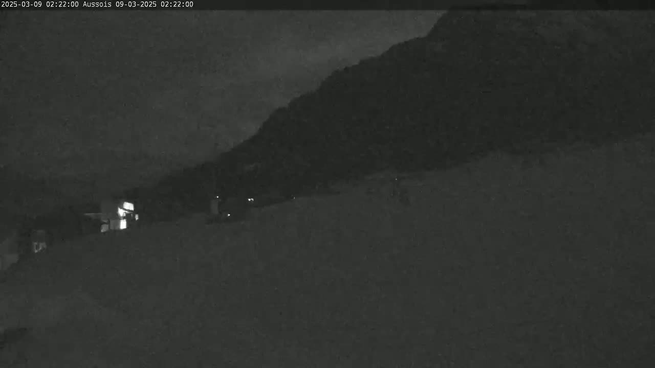 A nighttime, low-light view shows a dark hillside and a few faint lights in the lower left of the frame.