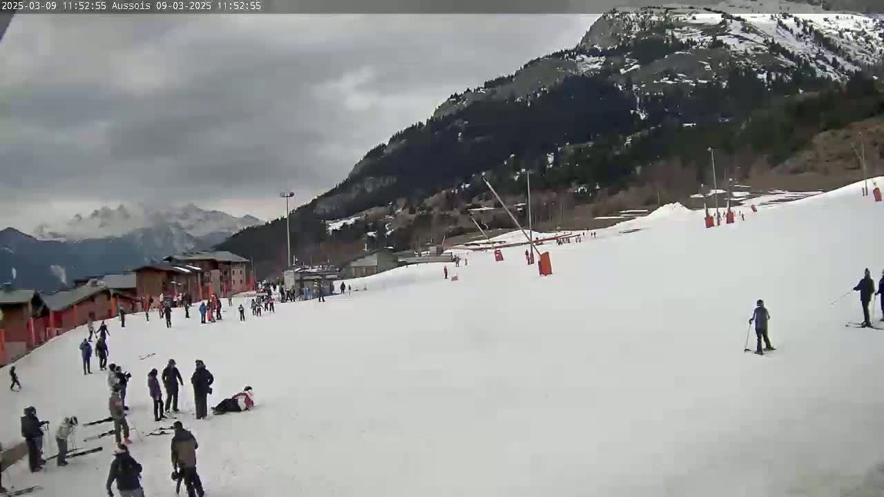 A snowy ski slope with several skiers and snowboarders, some buildings, and a mountain range in the background under a cloudy sky.