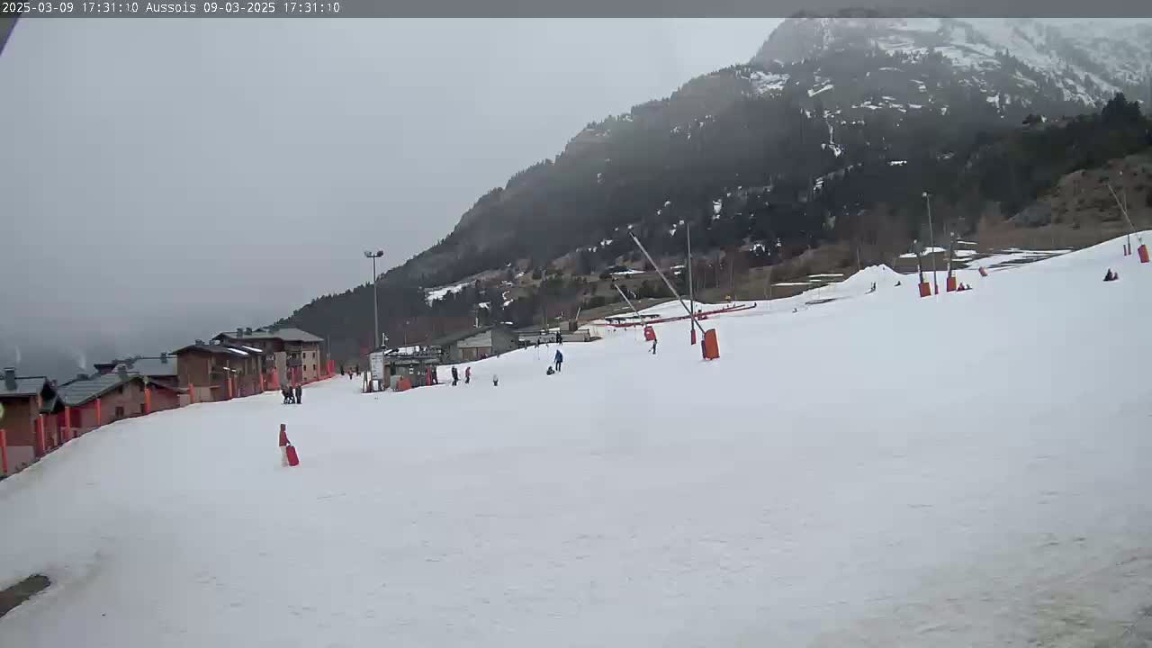 A snowy ski slope with several skiers and snowboarders, buildings, and a mountain in the background under an overcast sky.